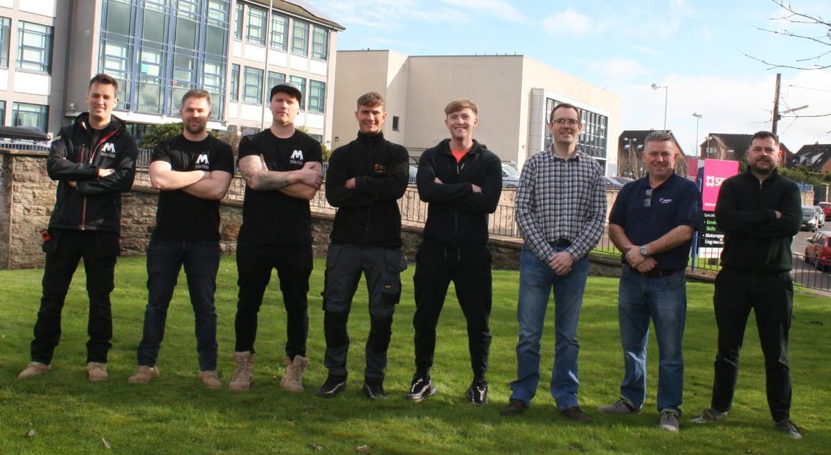 Eight men standing on grass area with college building in background